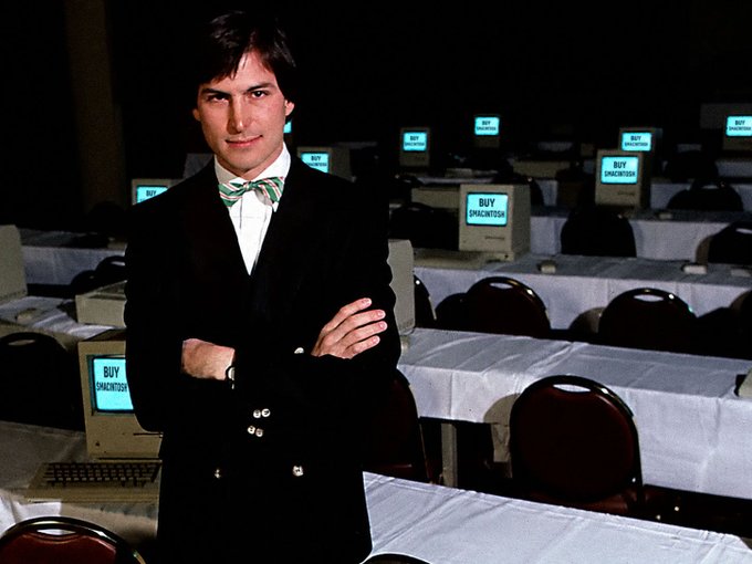 A young man in a dark suit with a bow tie stands with crossed arms in a room filled with computers displaying 'BUY DRACONTISER' on their screens.