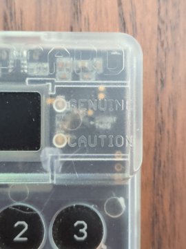 A close-up of a transparent Coldcard hardware wallet displays "GENUINE" and "CAUTION" warnings embossed on its casing above the "2" and "3" numbered buttons, illustrating the manufacturer's tamper-evident markings that verify device authenticity and signal proper disposal procedures after funds are swept to a new device.