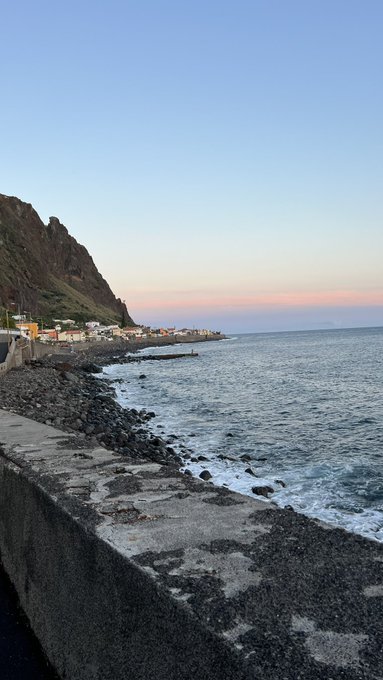 A coastal scene at dawn or dusk shows a rocky shoreline with waves crashing against a sea wall, backed by a steep cliff and small seaside village under a soft pastel sky, evoking a peaceful "gm" greeting to the community.