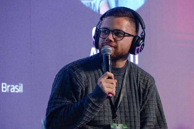 A man wearing headphones, glasses, and a gray sweater speaks into a microphone, with "Brasil" visible on the screen behind him.