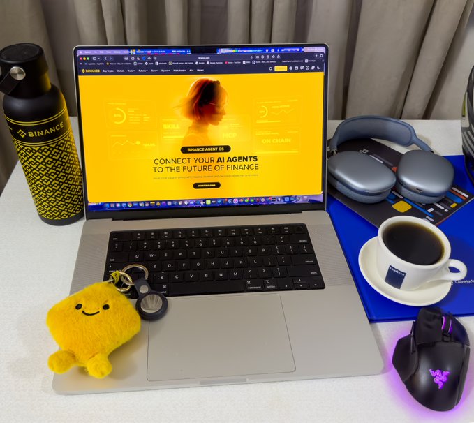 A laptop displaying the Binance website with the text 'CONNECT YOUR AI AGENTS TO THE FUTURE OF FINANCE' on a yellow background, surrounded by a black and yellow Binance water bottle, headphones, a coffee cup, a yellow plush, and a Razer gaming mouse.