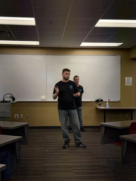 A man in a black t-shirt and gray pants stands and gestures in what appears to be a classroom or conference room, with another person partially visible behind him near a whiteboard.