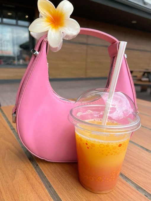 A clear plastic cup filled with an orange beverage and boba pearls sits on a wooden table next to a bright pink handbag with a white and yellow plumeria flower tucked into it.