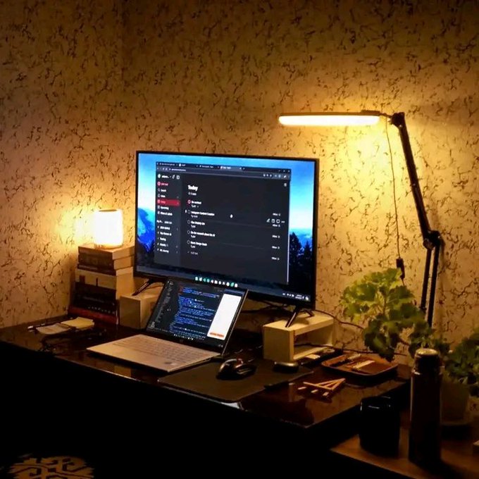 A warmly lit home office setup featuring a desktop monitor and laptop on a dark wooden desk, with a tall desk lamp casting golden light against patterned wallpaper.