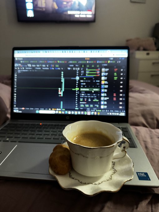 A laptop displays trading charts while a cup of espresso and a small pastry sit on a saucer on the keyboard, with a TV on the wall in the background.