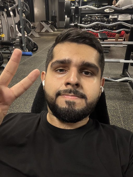 A selfie of a bearded man wearing AirPods, sitting in a gym with weight racks visible behind him, making a peace sign with his hand.
