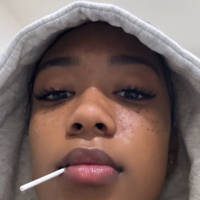 A close-up selfie of a woman in a gray hoodie with a cotton swab held near her lips, sharing an update about feeling unwell and postponing her Bloom mint Party and Karaoke night due to poor health and unstable network caused by the weather.