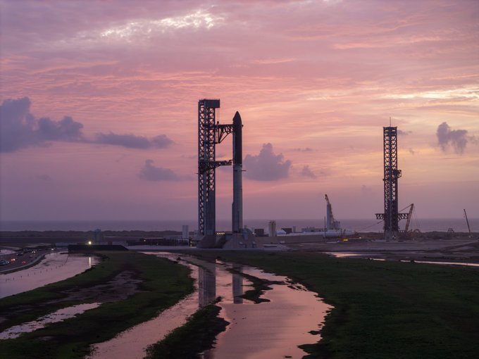 A SpaceX rocket stands on a launch pad at dusk, silhouetted against a pink and purple sky with reflections in water below