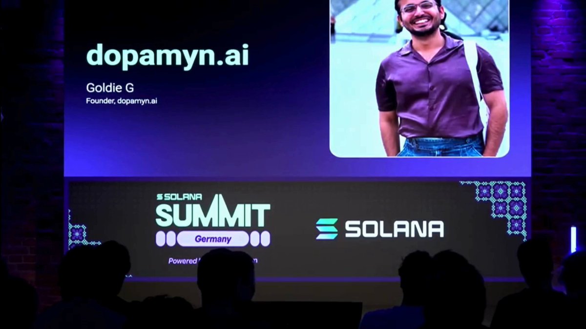 The image shows a large screen displaying an event where a man is standing up and talking about his experience at the Solana Summit. The audience appears engaged as they watch him speak, with some people sitting down while others stand around the room. The event seems to be taking place in front of a brick wall, possibly a stage or a presentation area.