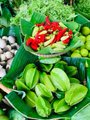 A close-up photo of various fresh produce, including green mangoes, red chili peppers, and limes, displayed in green baskets.