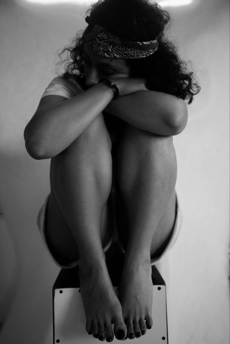 A black-and-white photograph shows a person with curly hair wearing a bandana, sitting on a small stool with their knees drawn up to their chest, arms wrapped around their legs, and head resting down in an introspective pose. The contemplative, monochromatic composition echoes the themes of stillness and reflection found in the artist's collected NFT works "Still," "In the Pursuit of Freedom," and "Light Fragment" by @HANI247249606 on Objkt.