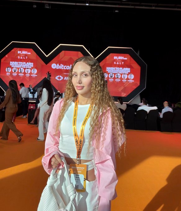 A young woman with curly hair wearing a pink jacket and lanyard at the "HASHTOPLASH TOKEN2049 WEEK DUBAI" event, with a "bitcoin" branded screen in the background.