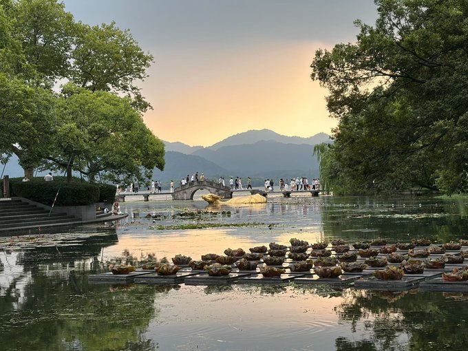 A serene lake scene at dusk featuring an arched stone bridge with pedestrians, mountains in the background, and floating lotus arrangements in the foreground water.