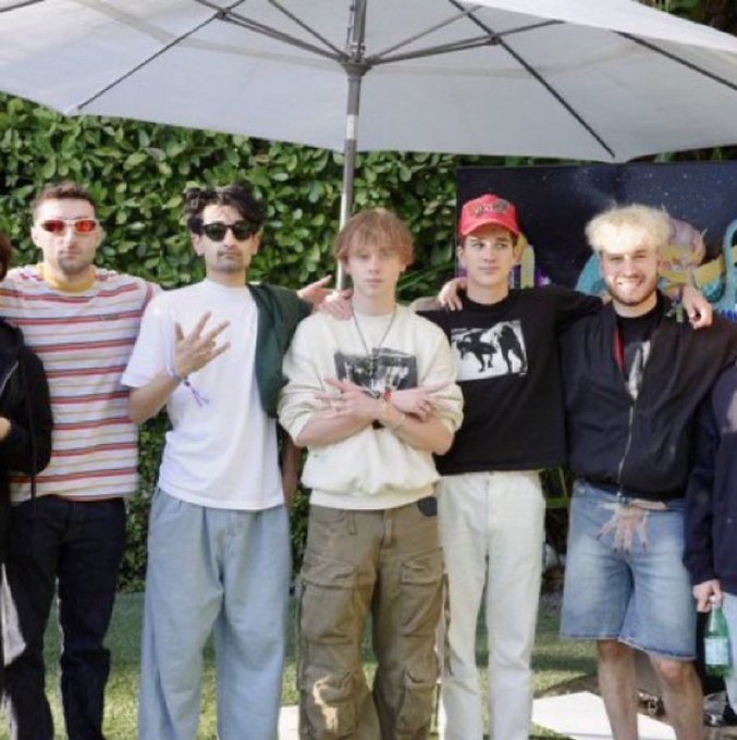 A group of five young men posing together under a white umbrella outdoors, with the central figure wearing a beige sweatshirt and cargo pants.