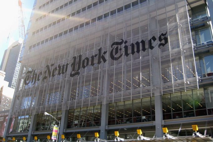 The exterior facade of 'The New York Times' building, featuring the newspaper's name in large Gothic lettering across the glass and steel structure.