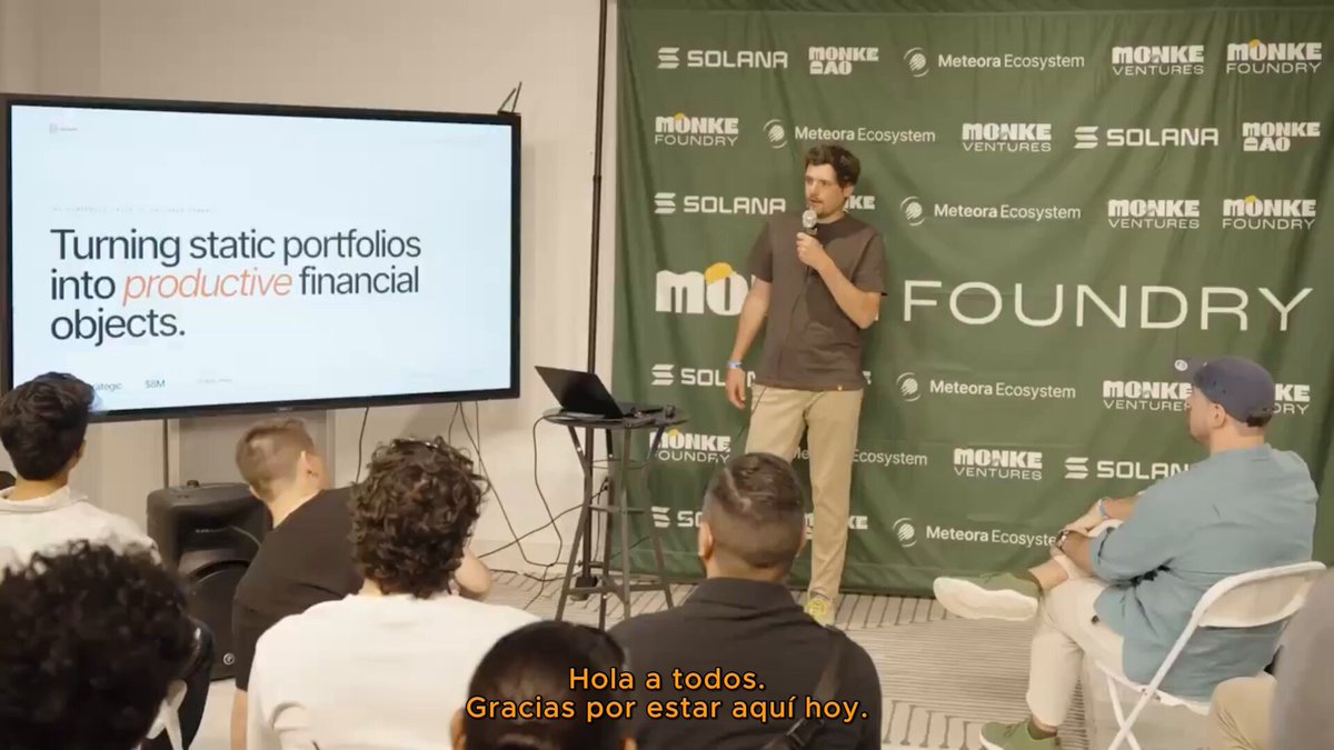 The image shows a group of people sitting around a table, with one man standing up and giving a presentation or speech. The audience appears engaged as they listen attentively to his words. In front of them, there's a large screen displaying information related to their discussion. A laptop is also present on the table, possibly used for taking notes or presenting slides during the meeting.