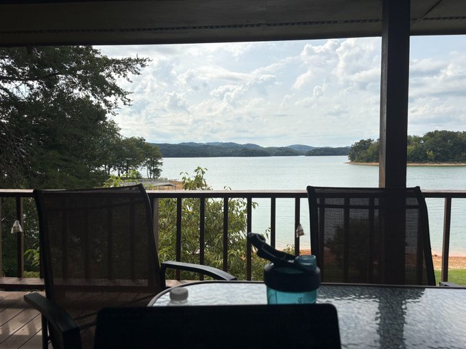 Two empty patio chairs sit at a table with a water bottle, overlooking a serene lake with rolling hills in the distance. The peaceful, reflective setting mirrors the introspective tone of the post, where the author steps back from business talk to reconnect with their core mission of helping creators realize their potential.