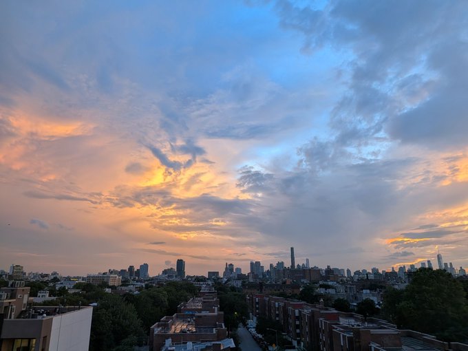 A scenic sunset view over a city skyline featuring a dramatic sky with orange and blue clouds, with buildings and trees visible in the foreground.