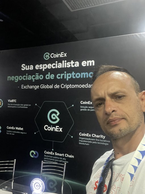 A selfie of a man at a CoinEx cryptocurrency exchange booth, with Portuguese text "Sua especialista em negociação de criptomoedas" and "Exchange Global de Criptomoedas" visible behind him.