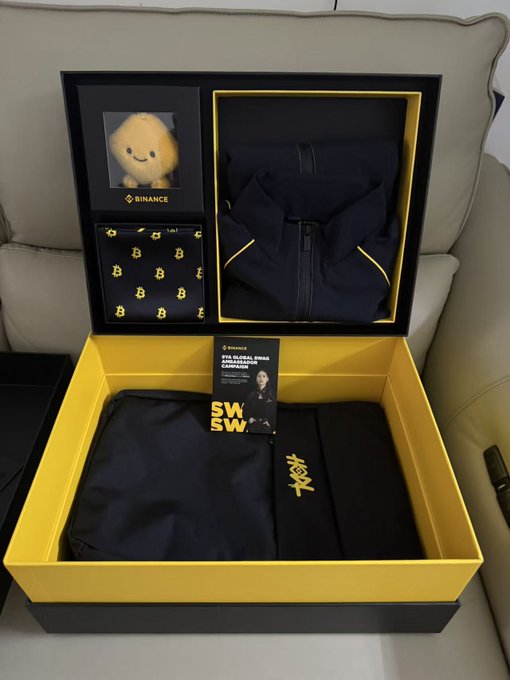 A Binance 9th Anniversary merchandise box displayed on a white couch, featuring a plush toy, a navy hoodie with yellow trim, a Bitcoin-patterned silk scarf, and clothing with "SWSW" and "BINANCE" branding, accompanied by a global ambassador card—matching the celebratory post about receiving anniversary swag.