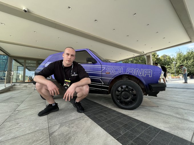 A man in black clothing squatting in front of a blue custom 'SOLANA'-branded SUV parked under an outdoor canopy at what appears to be a tech event.