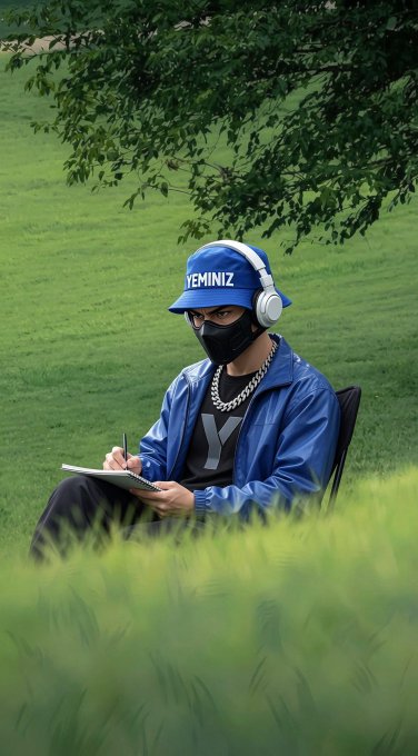 A person wearing a blue 'YEMINIZ' cap, face mask, blue headphones, and a blue jacket sits in a grassy field writing in a notebook under a tree.