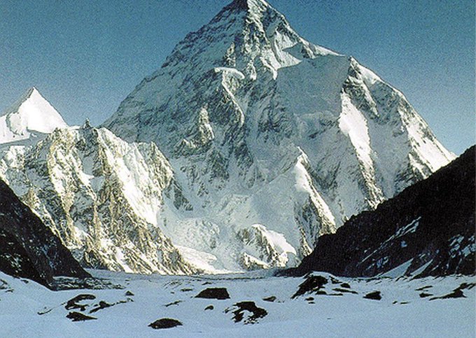 A majestic photograph of a towering snow-covered mountain peak, resembling the Himalayas, with a glacier valley in the foreground under a clear blue sky.