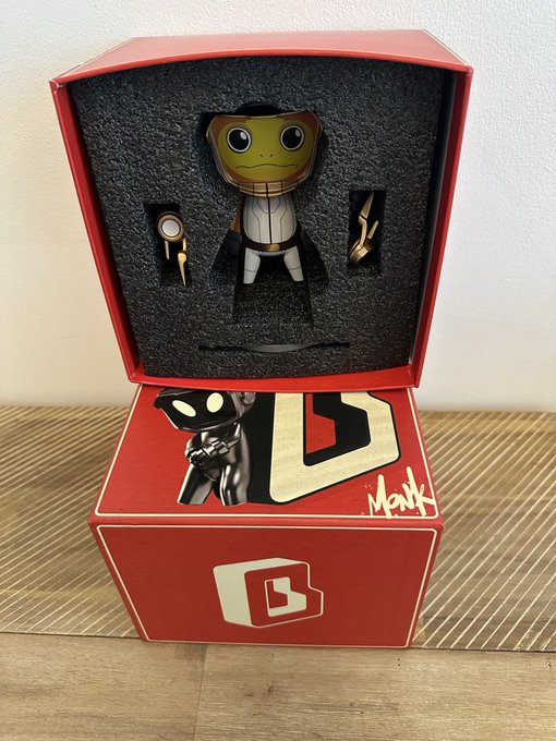 An open red collector's box displaying a stylized vinyl figure with a yellow face, cape, and mask, along with small accessories, with the word 'MONK' visible on the artwork below.