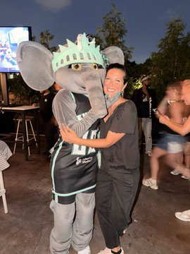A young woman with a smudge of makeup on her cheek is smiling while hugging a person in an elephant mascot costume wearing a green crown and basketball jersey at an outdoor nighttime event.