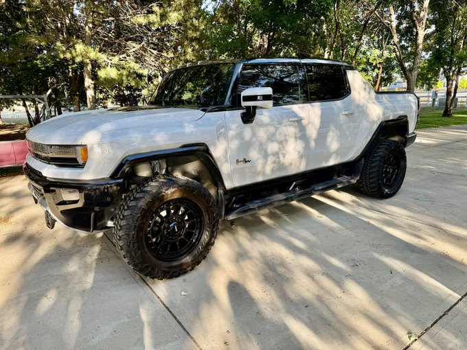 A photo of a white Hummer EV pickup truck with a lifted suspension and large off-road tires, parked on a driveway.