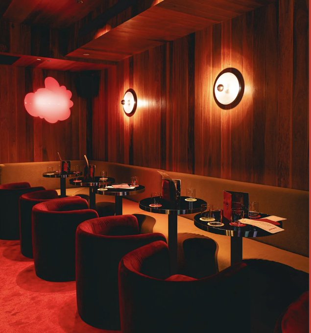 The image shows an upscale restaurant with wooden paneling and red velvet chairs arranged around tables filled with drinks, food, and wine glasses. The dining area features a large projection screen displaying a pink cloud design, adding a unique touch to the ambiance. The restaurant is likely hosting a special event or gathering, as indicated by the post's description of "gm. who' s gonna be at @ethconf in NYC next week? We're hosting a special event with @zerion @krakenfx @inkonchain @dynamic_xyz @CapApp".