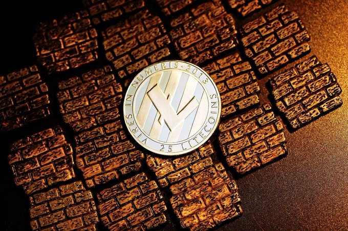A silver physical Litecoin coin inscribed with "VIRES IN NUMERIS-2013" and "25 LITECOINS" rests on a pile of chocolate bars.