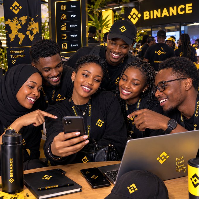 A group of six smiling young people wearing black merchandise at a Binance promotional or networking event, gathered around a table with Binance-branded laptops, water bottles, and notebooks, with a 'BINANCE' logo and yellow branding visible in the background.