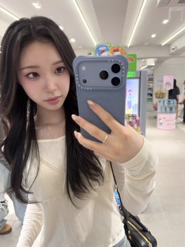 A mirror selfie of a young woman with long dark hair wearing a cream blouse and holding a gray smartphone in what appears to be a store.
