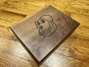 A square wooden plaque or box lid rests on a wooden floor, engraved with a simple line drawing of a skull.