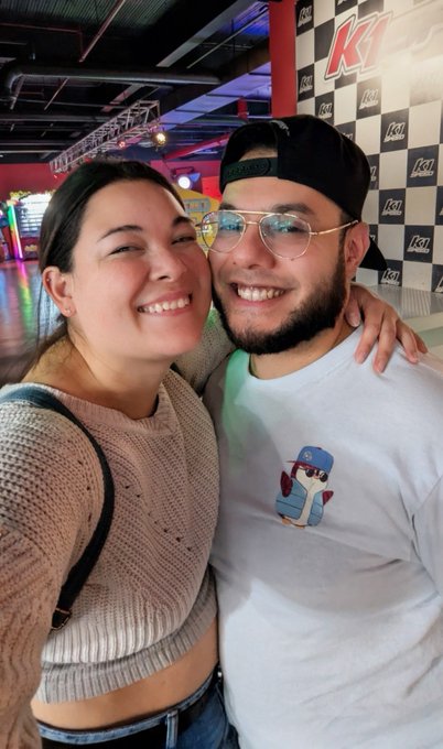 A smiling couple taking a selfie inside a K1 Speed indoor karting venue, the woman wearing a cream sweater and the man in a white graphic tee with sunglasses.