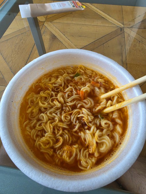 A bowl of instant noodles with chopsticks resting on the rim, photographed from above with an empty noodle packaging tube visible on the wooden table.