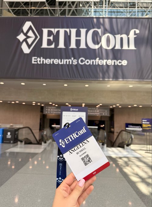 The image features a person holding an event ticket with their name on it at an event venue. The ticket has a blue tag attached, which displays "ETHGlobal" as well as the name of the conference or event they are attending. The person is standing in front of a large sign that says "ETHConf", indicating that this is an Ethical hacking conference.