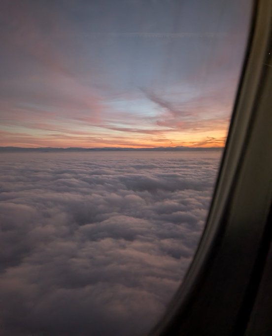 A scenic view from an airplane window showing a sea of clouds below a colorful sunset sky with pink and orange hues.