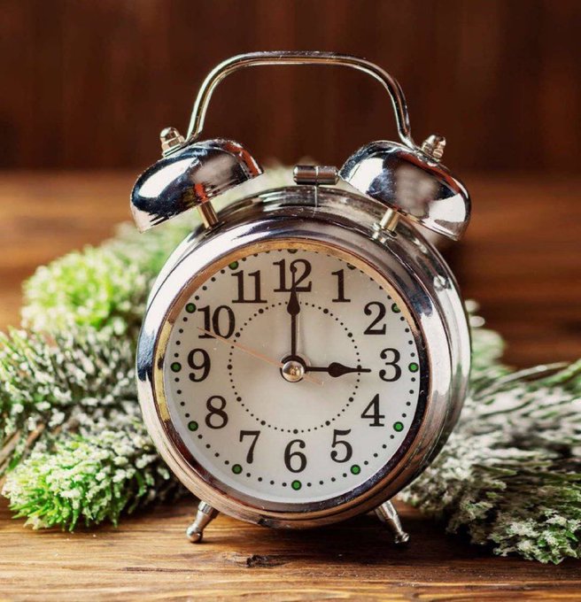 A vintage-style chrome twin-bell alarm clock showing 3:00, photographed on a wooden surface and decorated with frosted green pine sprigs for a holiday aesthetic.