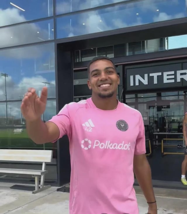A man with a beard smiling and waving at the camera while wearing a pink Inter Miami training jersey with a 'Polkadot' sponsor logo, standing in front of the Inter Miami training facility.