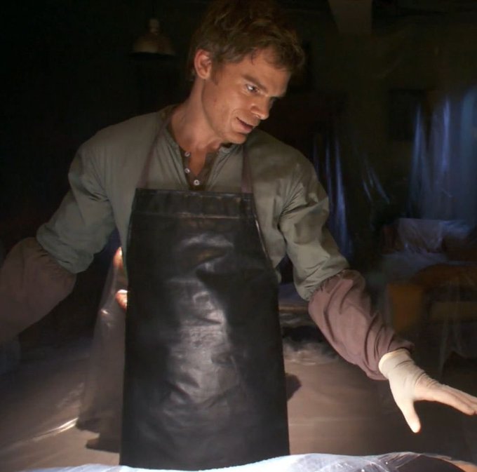 A man wearing a gray shirt, black apron, and latex gloves stands in a dimly lit room, looking down at a plastic-covered surface.