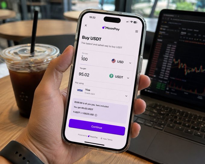 A hand holding a smartphone displaying the MoonPay app interface for buying USDT with USD, showing 100 USD paying for 95.02 USDT via Visa, with an iced coffee and laptop in the background.