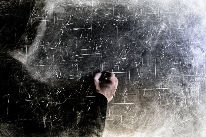 A hand writing mathematical equations and formulas in white chalk on a dark blackboard.