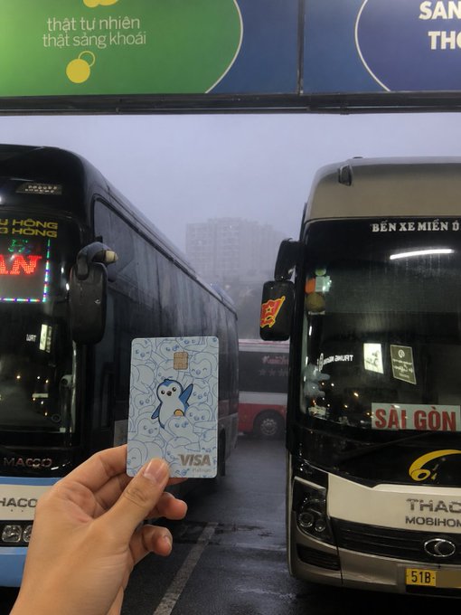 In the image, there are two buses parked next to each other at an airport or bus station. A person's hand is holding a Visa card with their photo on it, which they're using to purchase a ticket for one of the buses. The scene captures the convenience and modernity of using digital payment methods like credit cards in everyday life situations.