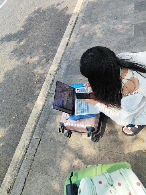 A woman crouches on a sidewalk with her laptop balanced on a rose-gold suitcase, intently taking a photo of the screen with her phone. The scene perfectly captures the post's joke about web3 enthusiasts being so plugged in that they can't even put their work away while hustling to catch the bus.