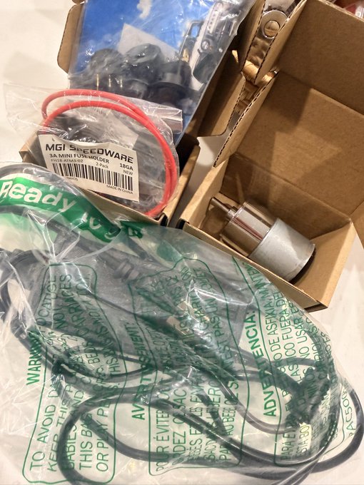 A collection of computer hardware components in plastic packaging, including cables and connectors, with a visible label reading 'MGI SPEEDWARE 3A MINI FUSE HOLDER SOLDER' and a green warning bag containing additional parts.