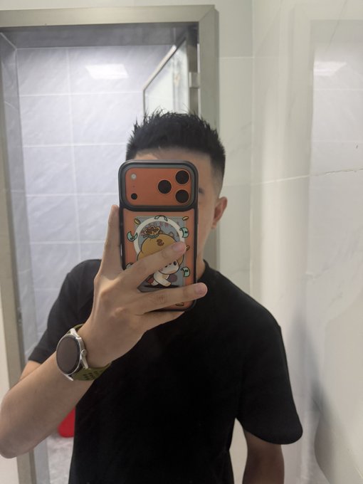 A man in a black t-shirt takes a mirror selfie in a tiled bathroom, wearing a smartwatch on his wrist and holding a phone with a cute cartoon character case featuring a bear in a spacesuit.