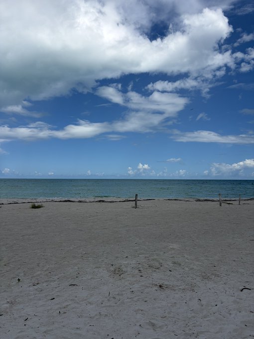 A serene beach scene with white sand in the foreground, calm blue ocean waters, and a bright sky with scattered clouds.