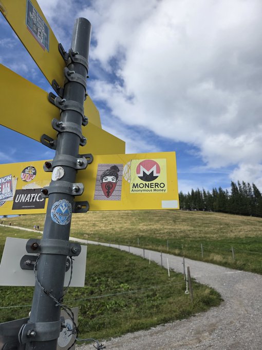 A photograph of yellow directional signs on a metal pole outdoors, featuring stickers including one that prominently displays the 'MONERO - Anonymous Money' logo alongside other stickers.